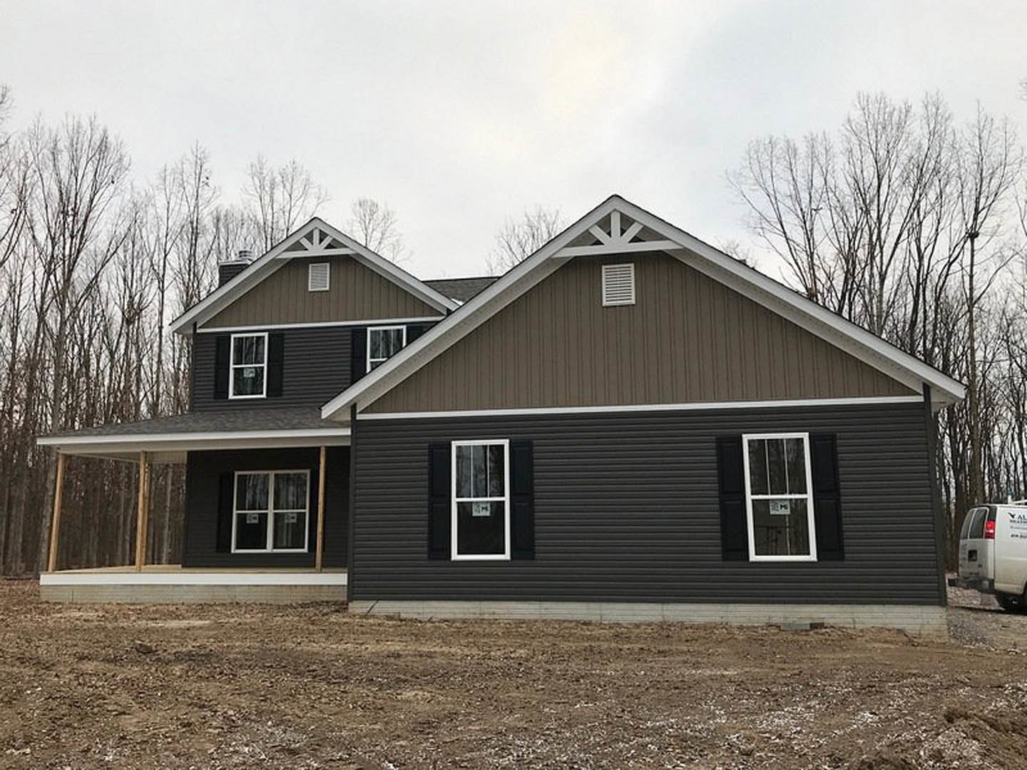 Two-story house with light siding, gabled roof, white-framed windows, surrounded by bare dirt and mature trees; white van parked near the back, wall vent visible.