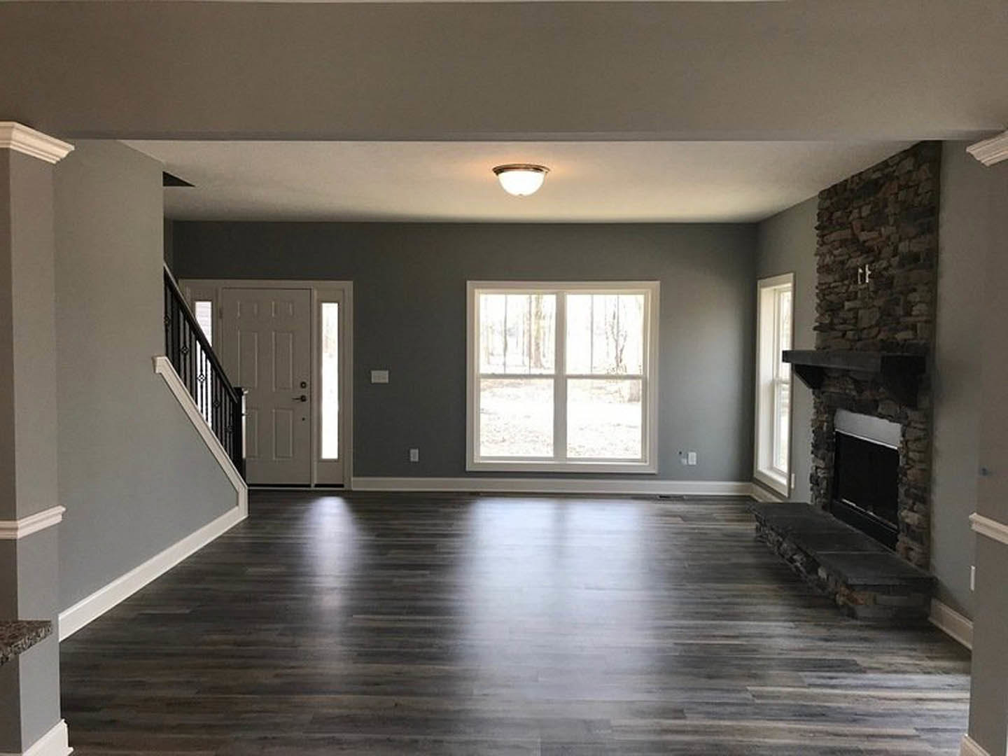 Living room with stone fireplace and ledge, hardwood floor illuminated by natural light, large window overlooking forest, white door with glass panels, plaster walls and ceiling