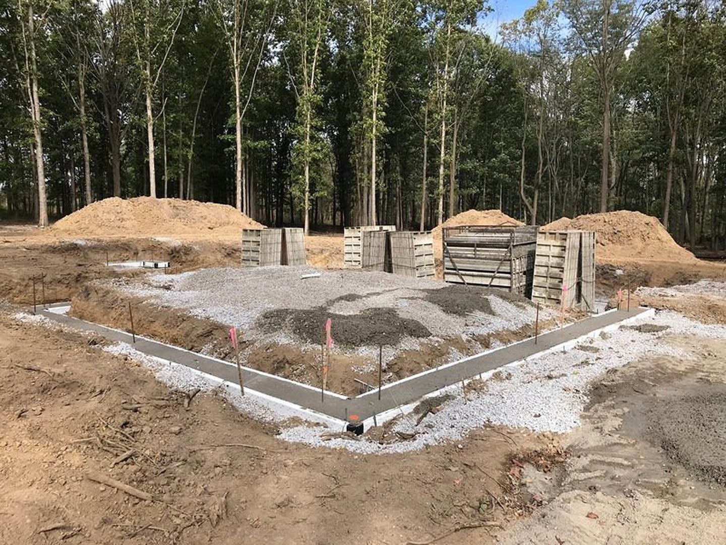 Foundation construction site with exposed soil, wooden crates, concrete slabs, and metal structure, bordered by trees under open sky