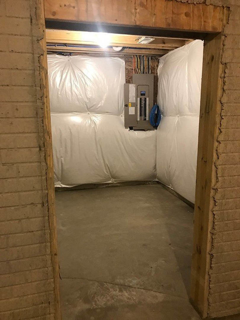 Unfinished basement room with concrete floor, white plaster walls, open door, ceiling light, and white plastic covering draped over a chair.