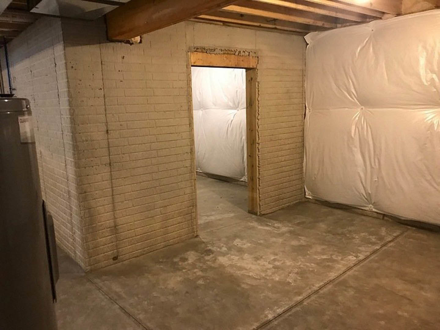 Exposed brick wall with wooden beam, white plaster ceiling, concrete floor, and wooden door in a basement room