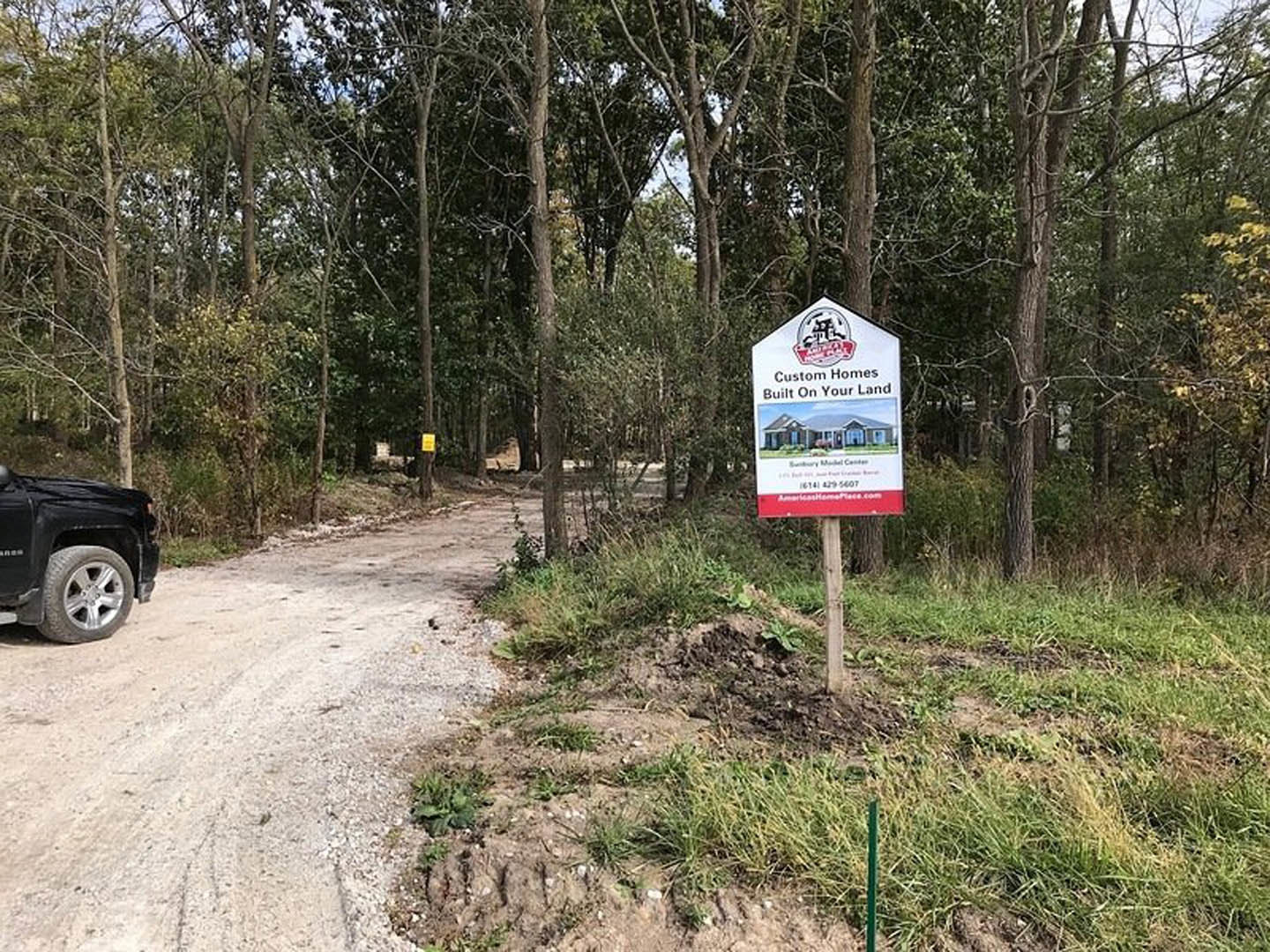 Black truck parked on a dirt road beside a grassy area with a sign displaying a house illustration, surrounded by trees under a clear sky.