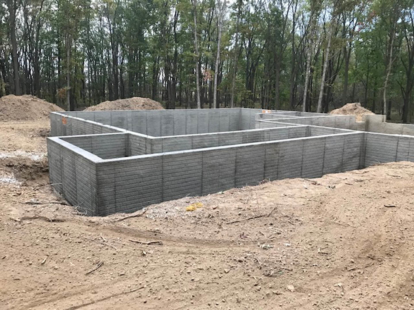 Concrete foundation slab surrounded by soil and construction materials, with trees and plants in the background under a partly cloudy sky
