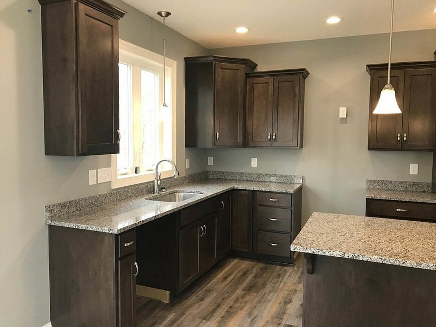 Kitchen with dark wood cabinets, black and white granite countertops, stainless steel faucet, light fixture above door, sunlight streaming through window, white utensil on wooden