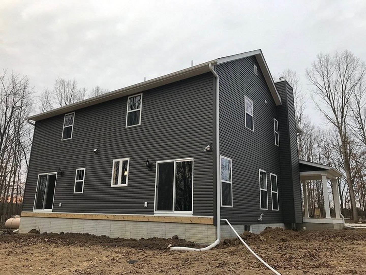 White pillared porch with screen door, unfinished siding, several windows, leafless tree, exposed pipe in dirt, construction materials scattered around yard