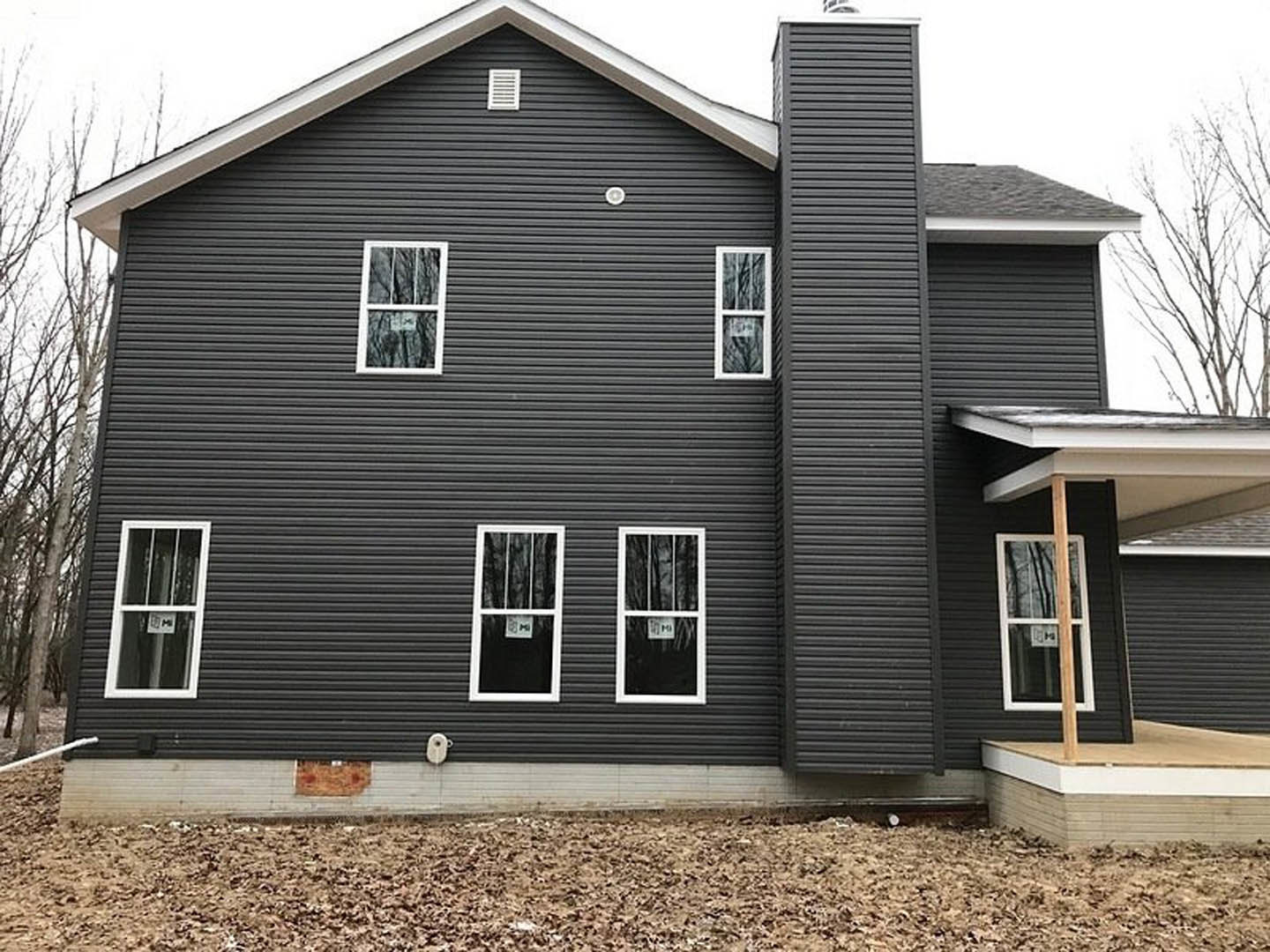 Black siding house with multiple white-framed windows, prominent brick chimney, and front door, surrounded by outdoor landscaping.