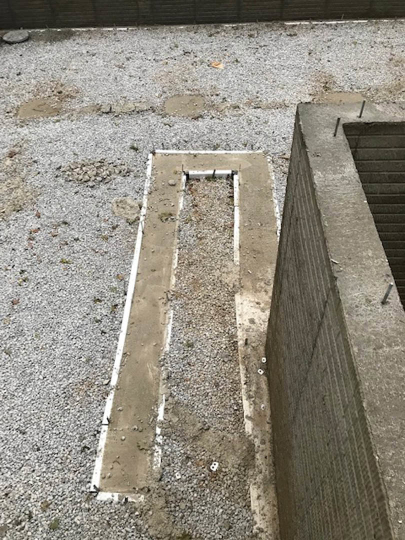 Concrete drainage channel set into gravel ground, bordered by a concrete retaining wall with a rectangular drain opening.