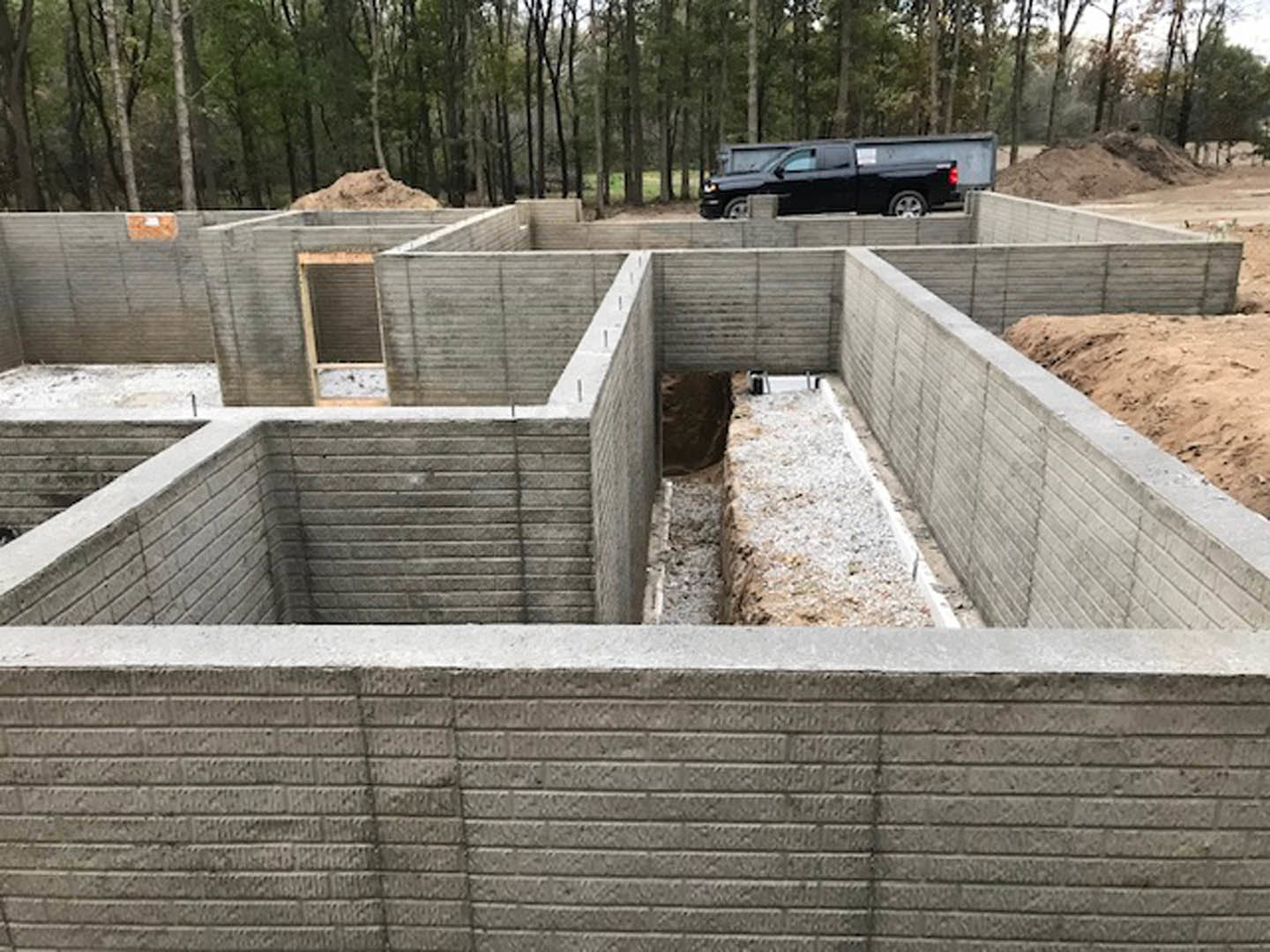 Partially constructed custom home with exposed concrete foundation, composite exterior walls, and black pickup truck parked in the background; surrounding area includes trees