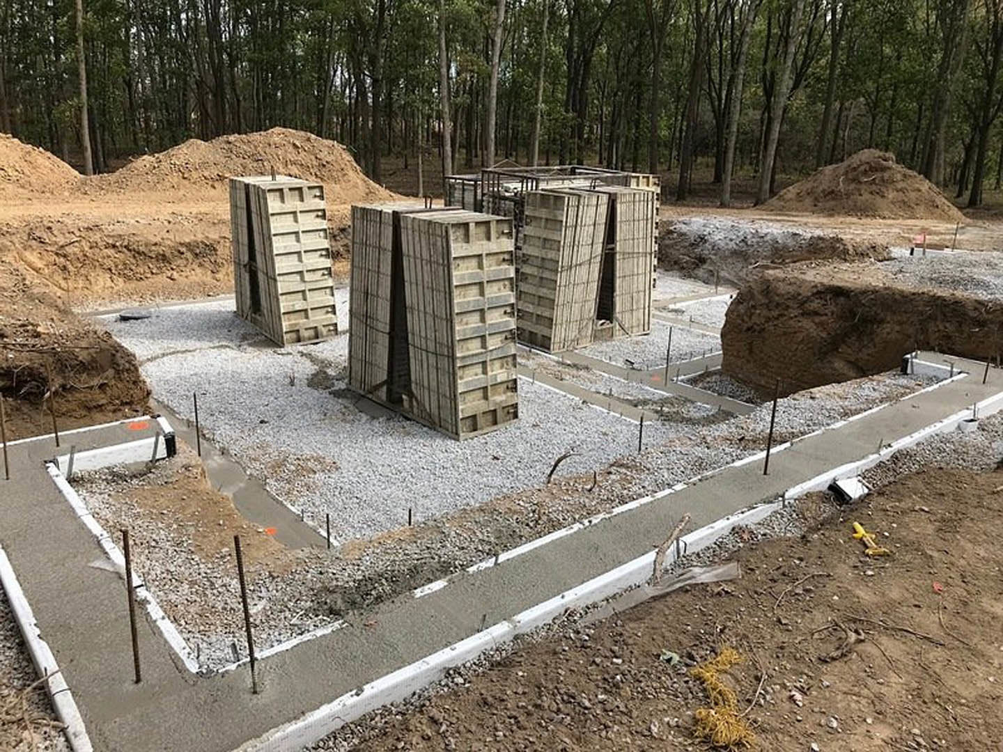 Concrete foundation walls and slab at a residential construction site, surrounded by soil, trees, and unfinished ground.