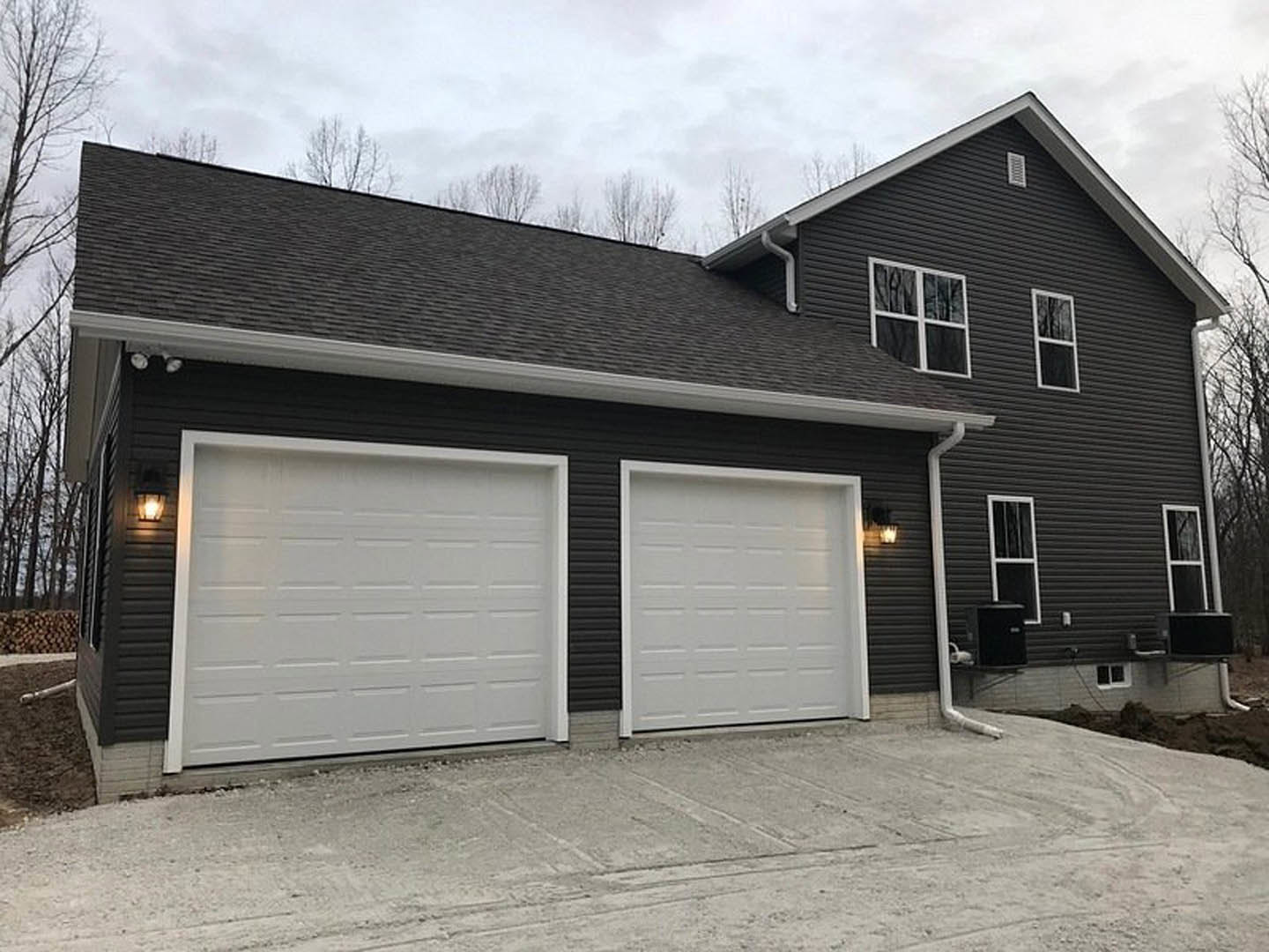 Two-car garage with white doors and black siding, concrete driveway, white-framed window, Plattsburgh Air Force Base visible in background, trees and sky surrounding property