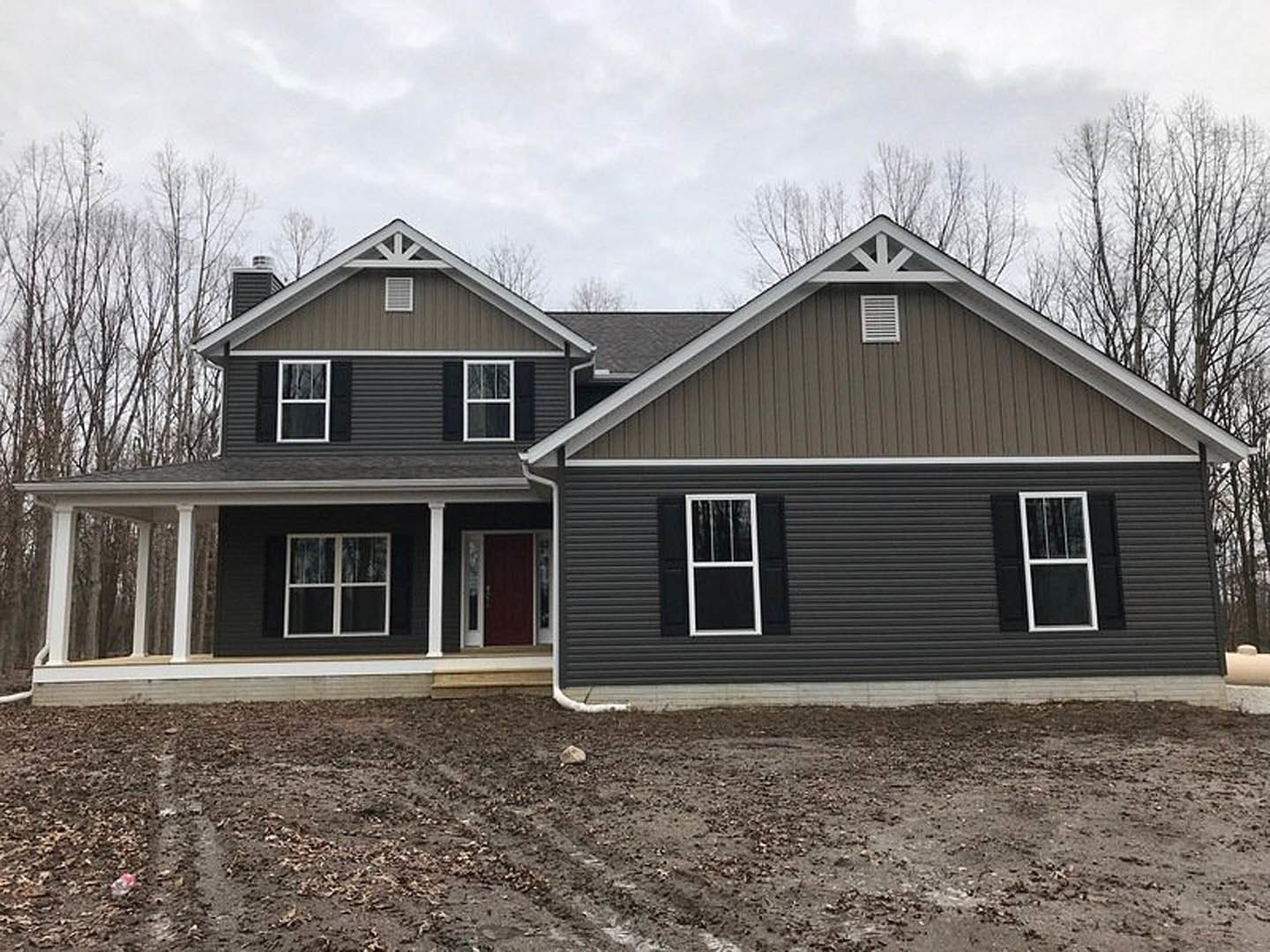 Two-story house with black siding, red front door, white-framed windows, and a dirt driveway bordered by sparse trees