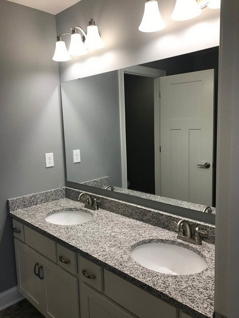 Bathroom with a wide mirror above a white countertop featuring black speckles, chrome faucet, three-bulb light fixture, and modern cabinet.