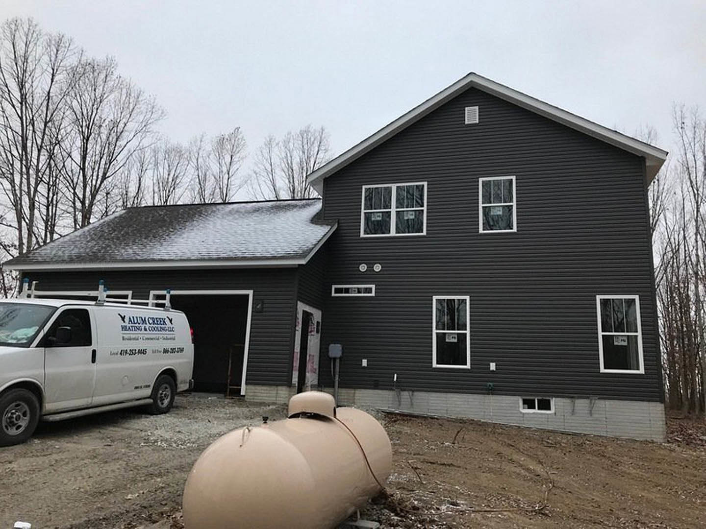 Two-story house with tan siding, attached garage, white-framed windows, large round tan tank beside driveway, white van with blue and black lettering parked in front, tree and car