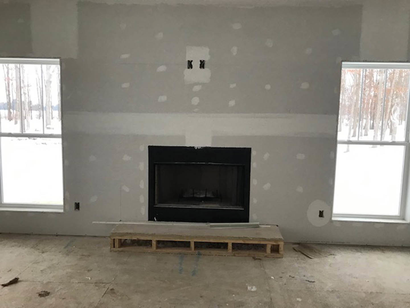 Modern living room featuring a black fireplace with active flames, large windows framing a snowy landscape, light plaster walls, and hardwood floors.