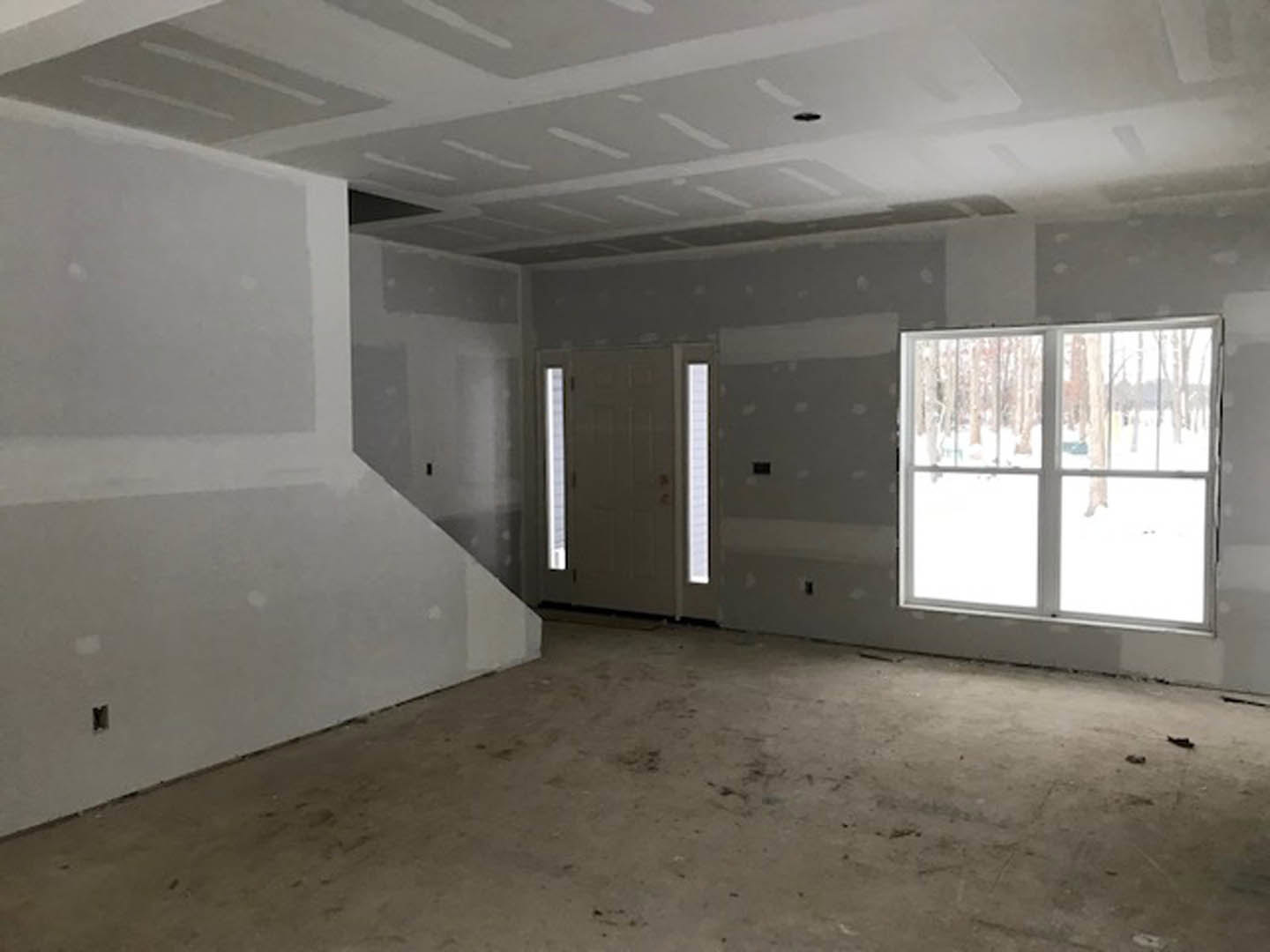 Open room featuring a white staircase against a white wall, large windows revealing snowy landscape, white door with glass panel, dirty floor, and ceiling with white linear