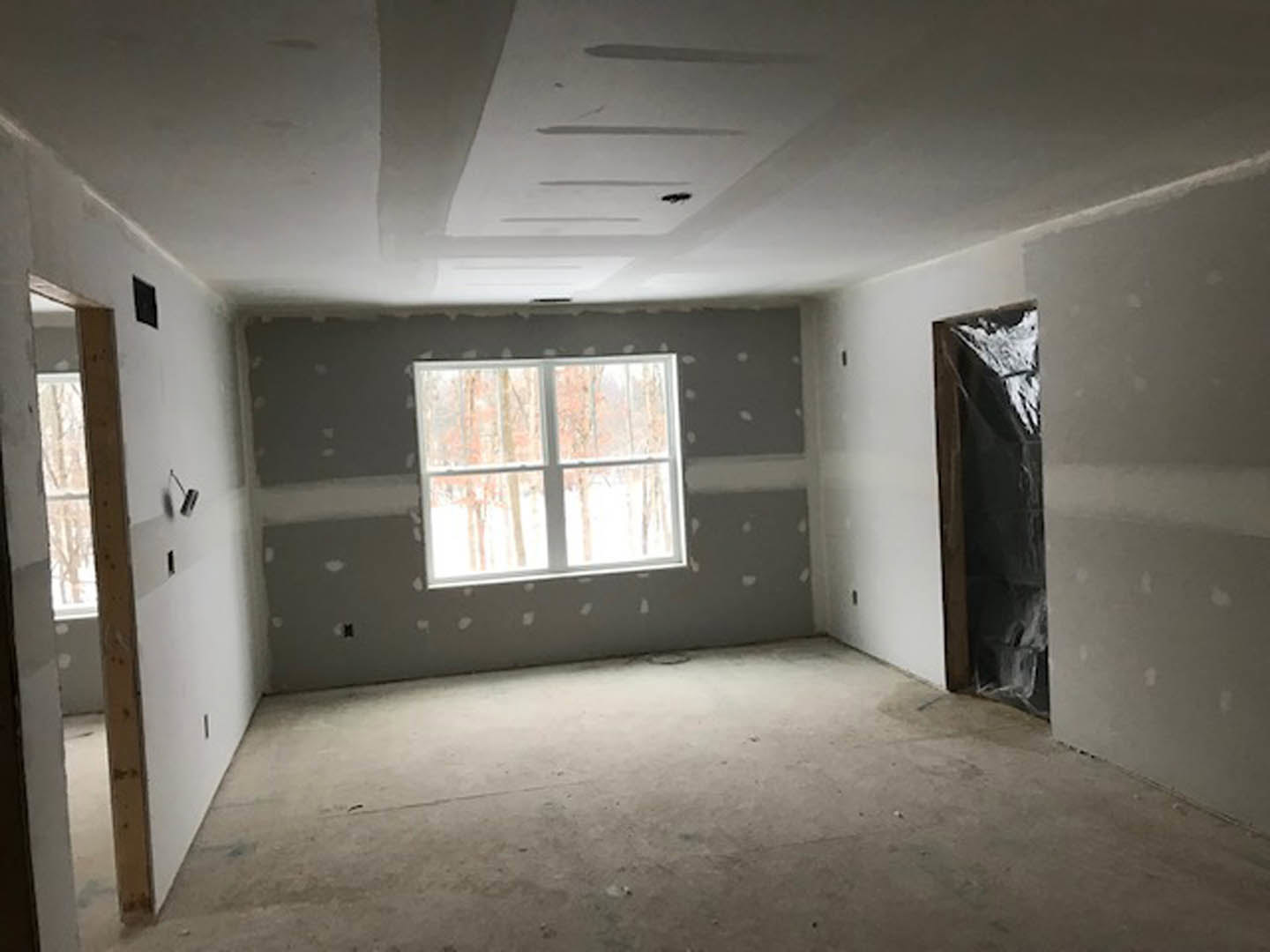 Sunlit room with plaster walls, large window framing leafy trees, concrete floor, black rectangular fixture with brown trim, and a wooden door.