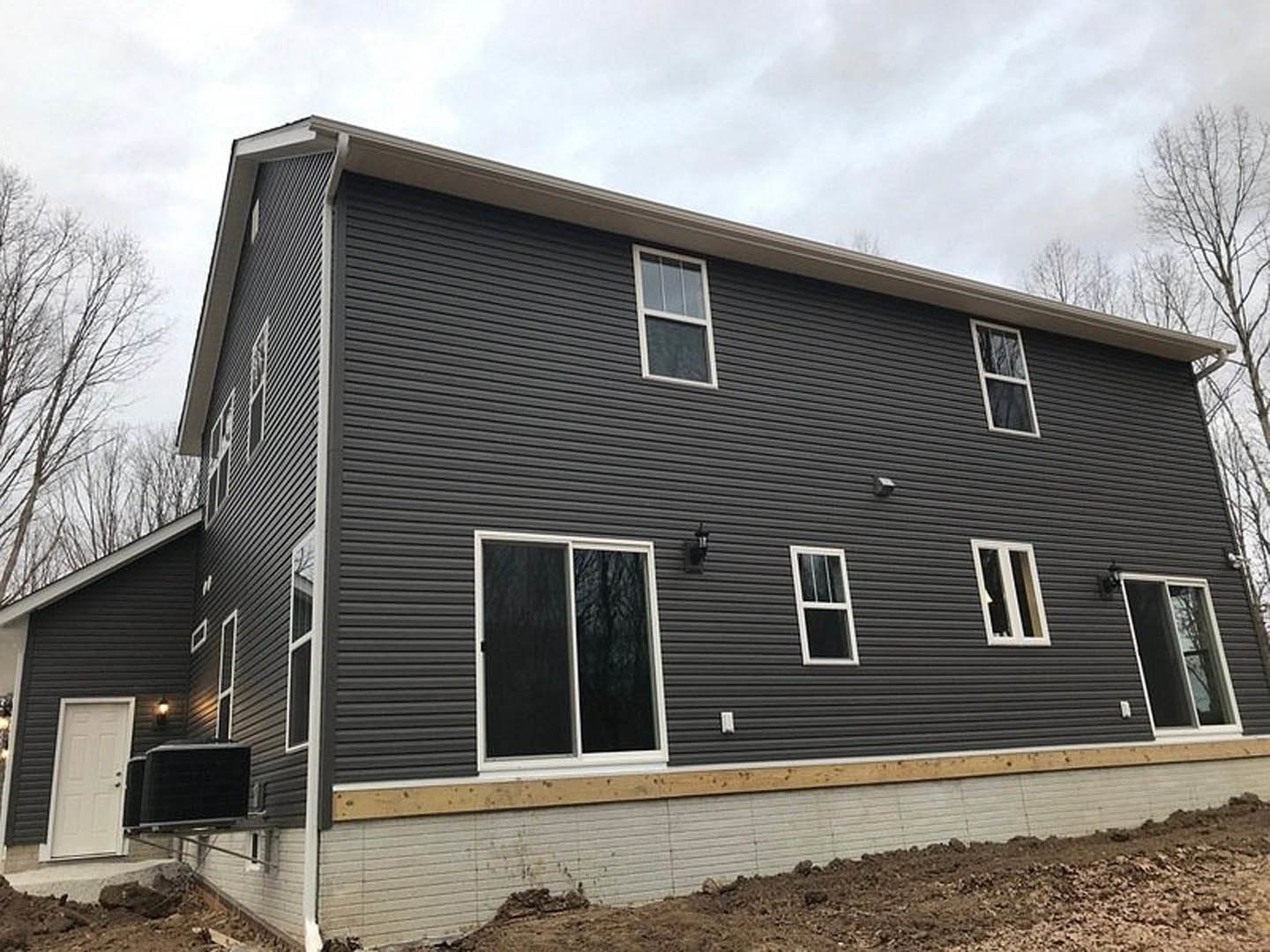 Two-story house under construction with exposed framing, white window frames, white door, black siding, and large dirt field in foreground; tree visible near building.