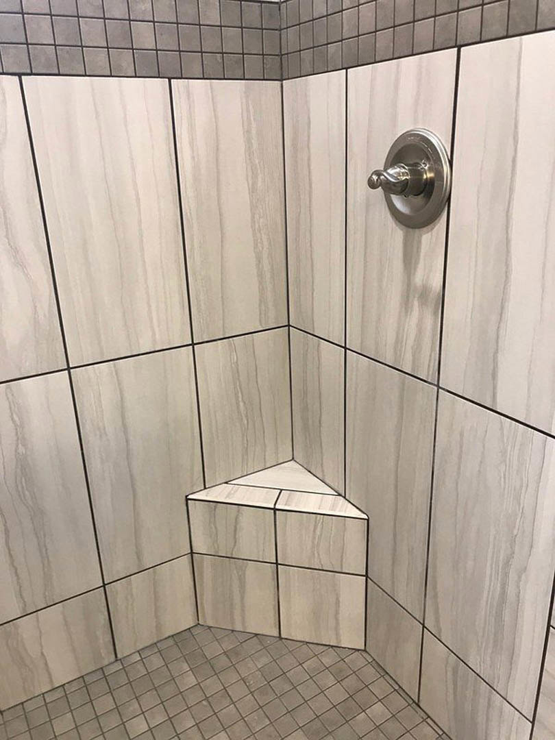 Modern bathroom shower with chrome shower head, large rectangular wall tiles, and light gray tile flooring; includes a close-up of a metal hook, tile corner detail, white