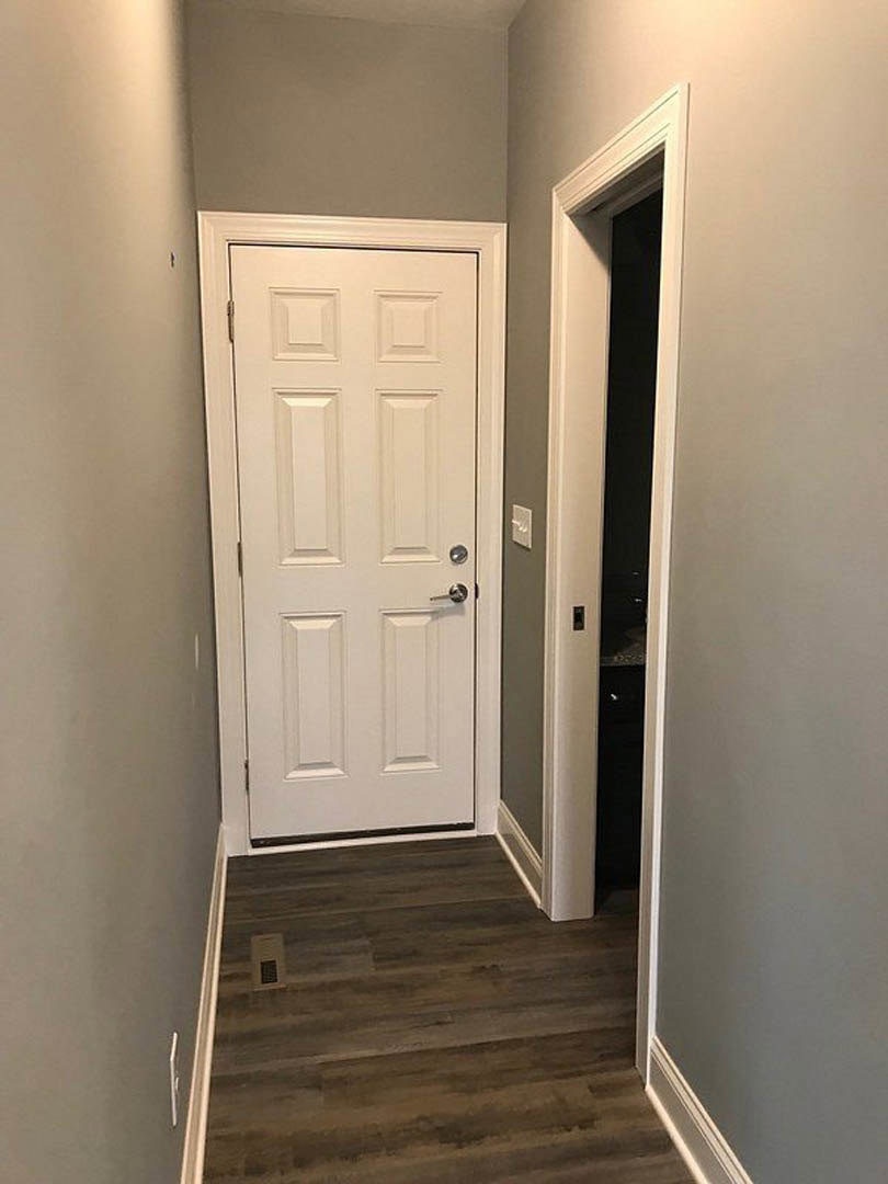 White door with silver handle in hallway, wood floor with vent, white walls, black rectangular object near doorway