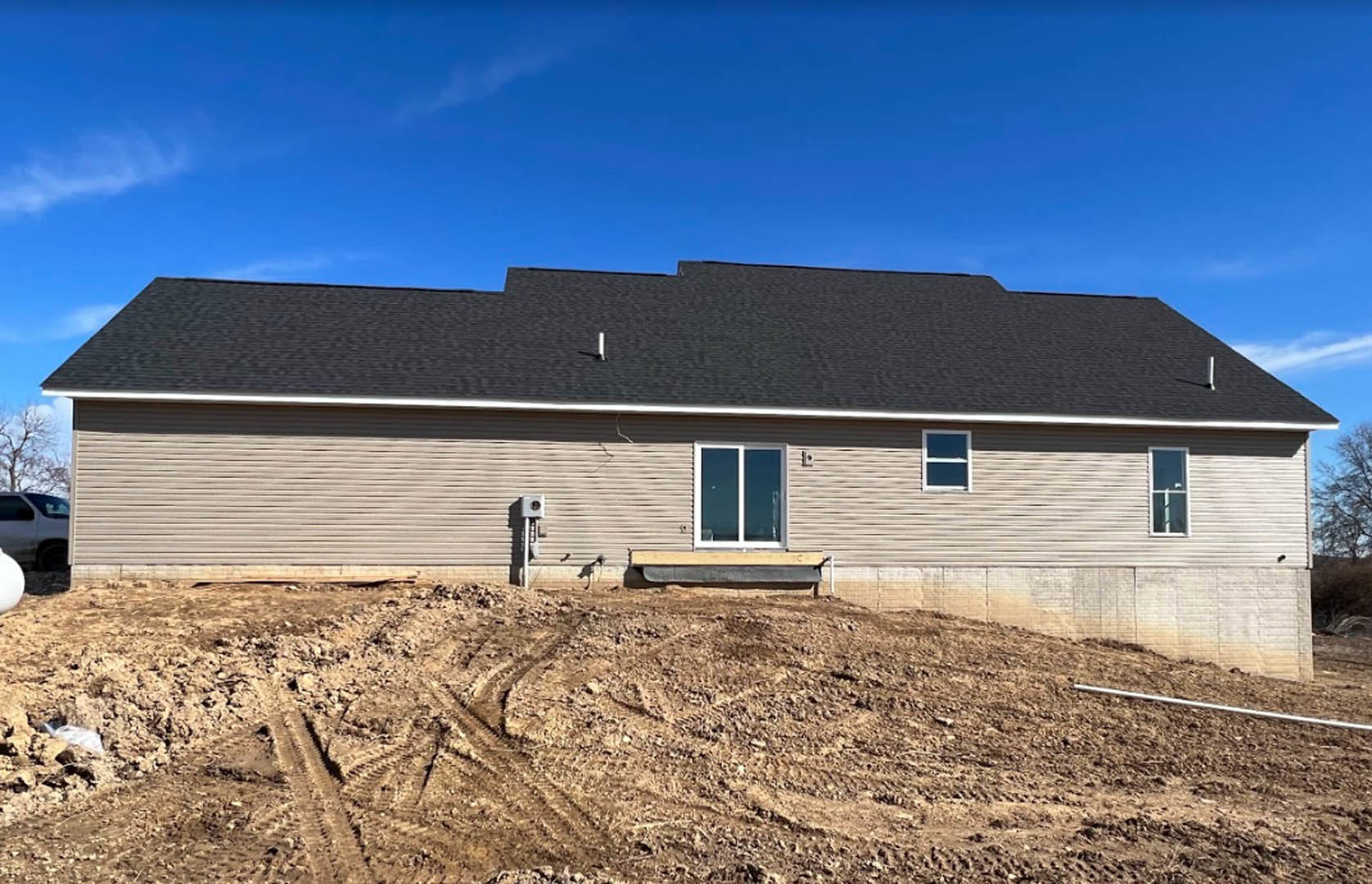 Light-colored siding house with white-framed windows, bench near entrance, dirt lot with tire tracks in foreground, cloudy sky overhead