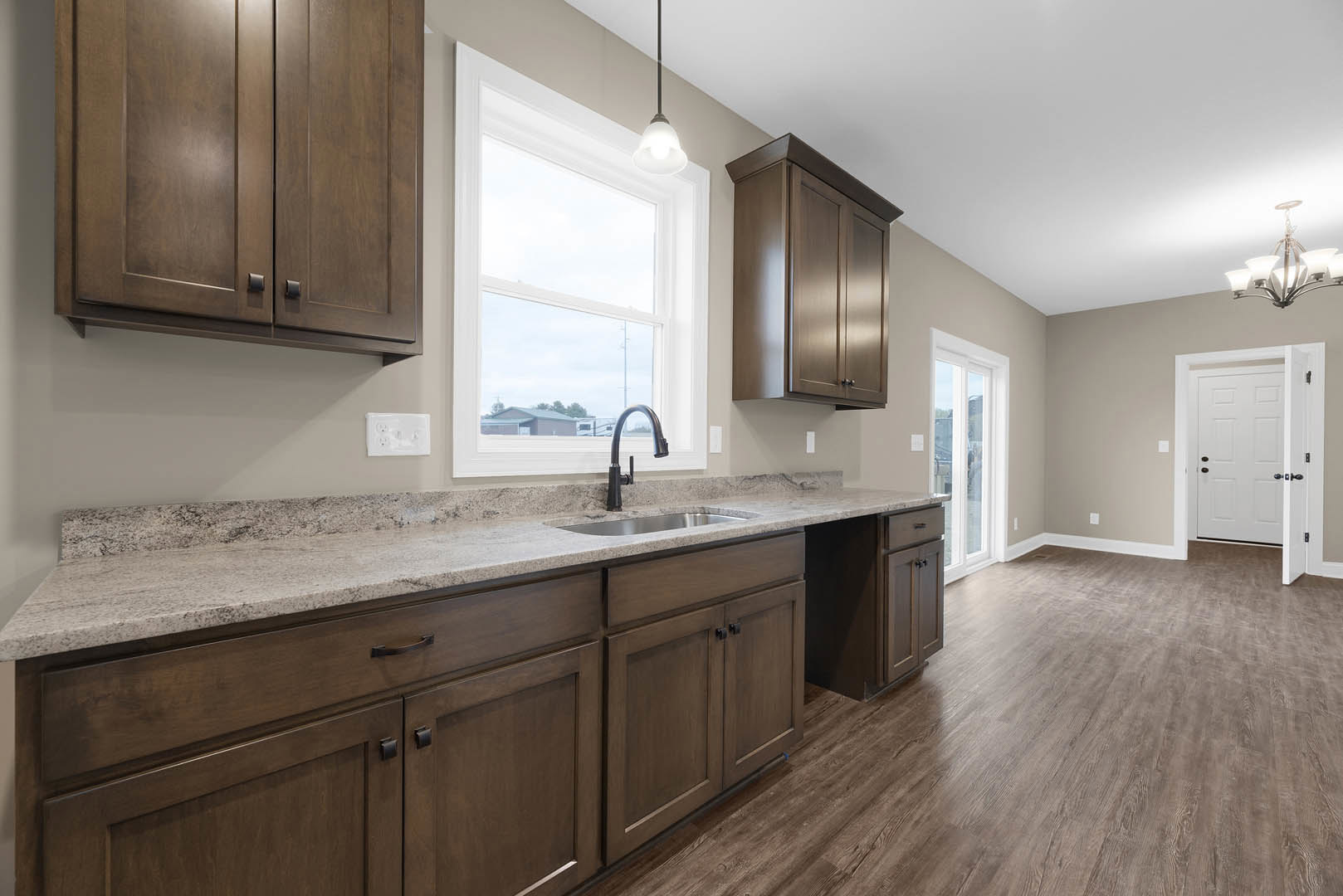 Kitchen with dark wood cabinets, stainless steel sink, tile backsplash, white electrical outlet, black cabinet handles, modern chandelier, light fixture above window, white door