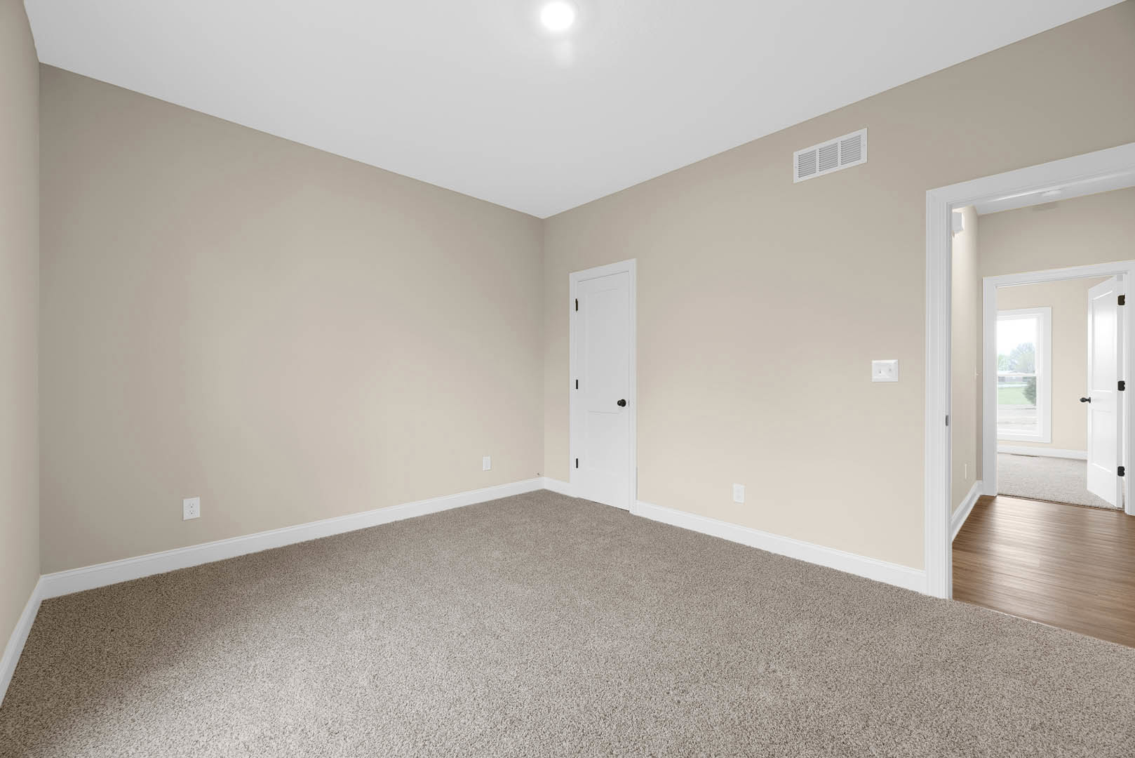 Beige walls and light carpet, white door with black knob, wood floor transition, wall vent, white ceiling with circular fixture