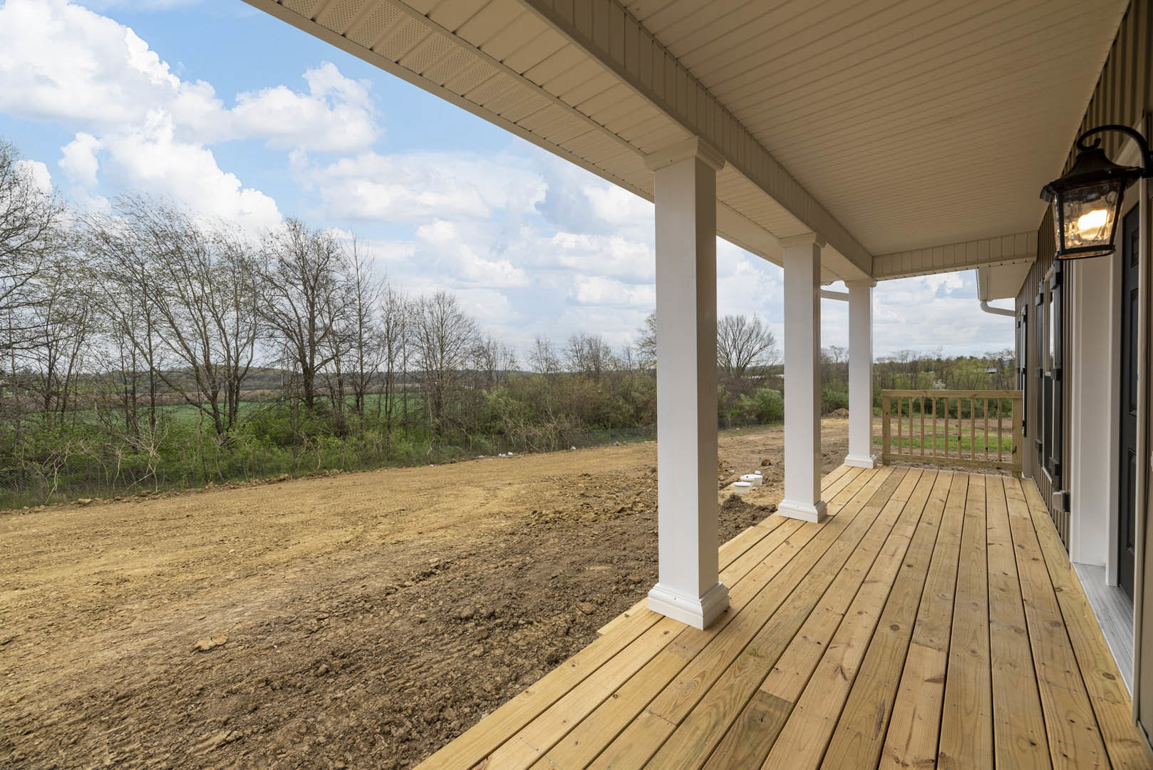 Covered porch with white pillars, wooden deck, black lamp fixture, and dirt field surrounded by grass and trees under a cloudy sky