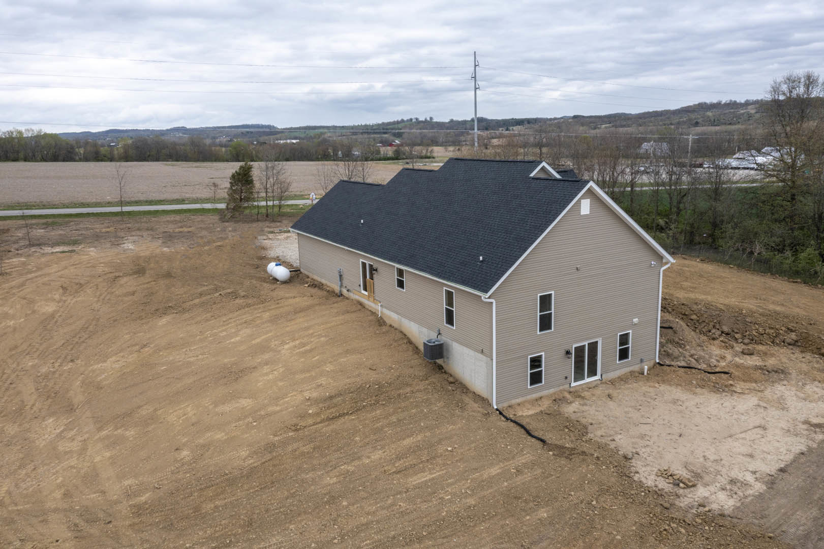 Modern house under construction with exposed framing and roof, surrounded by a dirt field and scattered green trees beneath a cloudy sky.