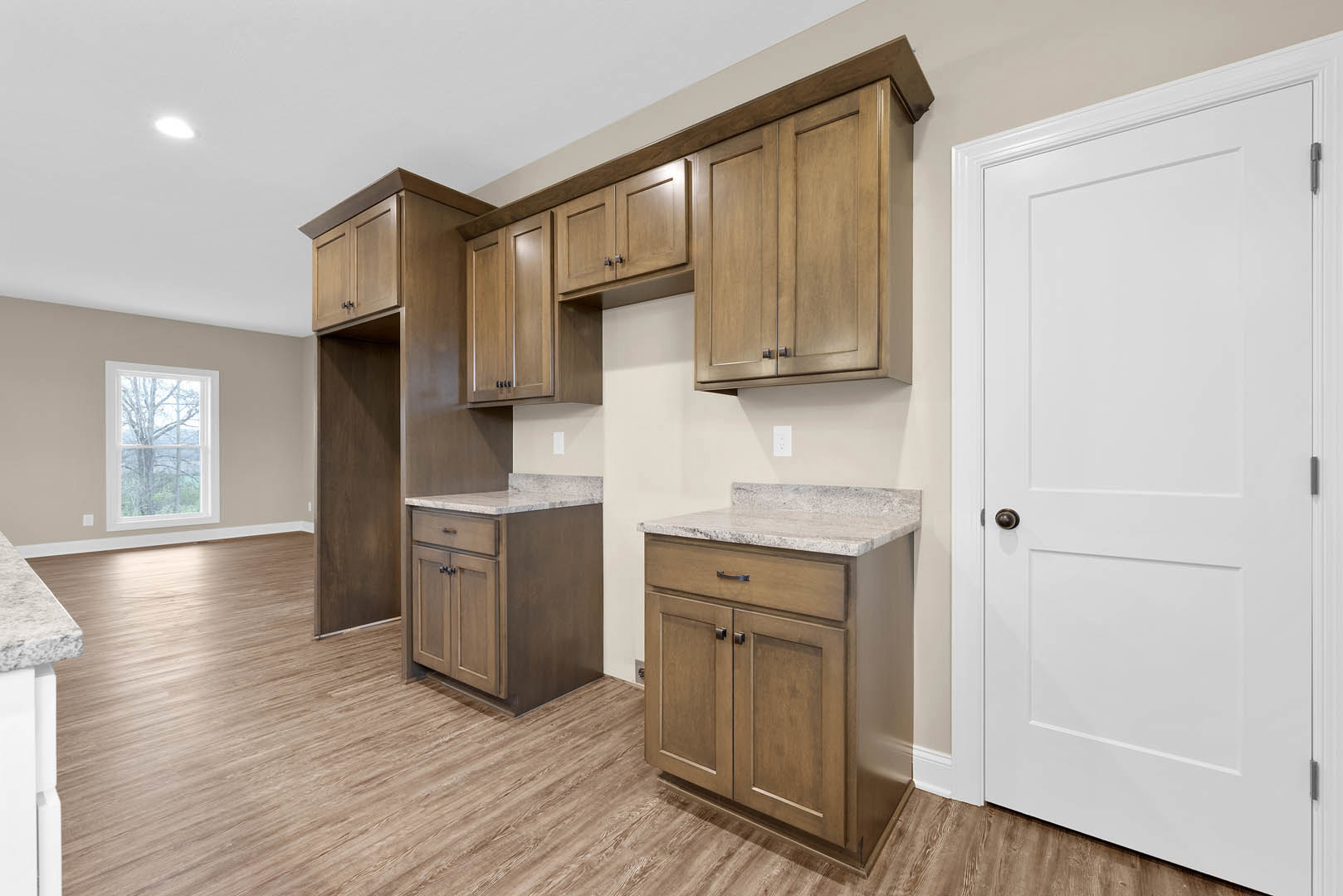 Kitchen with wooden cabinets, marble countertops, white door with bronze handle, window overlooking trees, light-colored flooring, and built-in appliances
