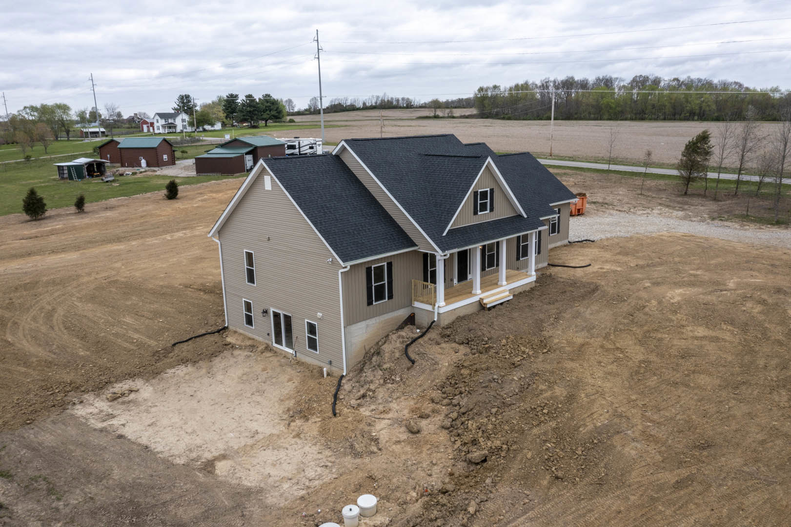Wood-framed house under construction on dirt lot with scattered building materials, surrounded by open field and trees under cloudy sky