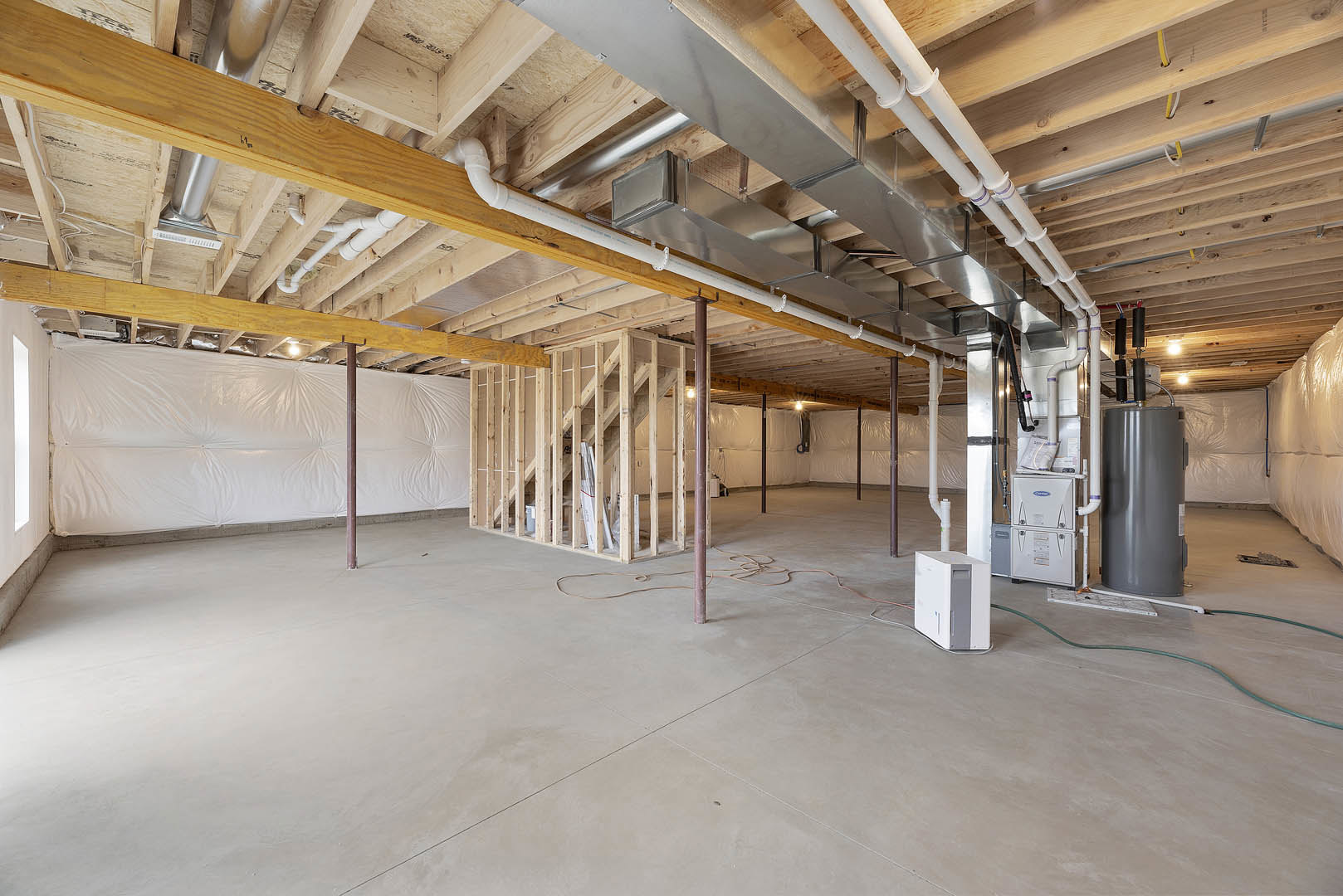 Unfinished basement with exposed steel pipes, wood framing, concrete floor, white plastic wall covering, grey cylinder with white label, white rectangular object with grey handle