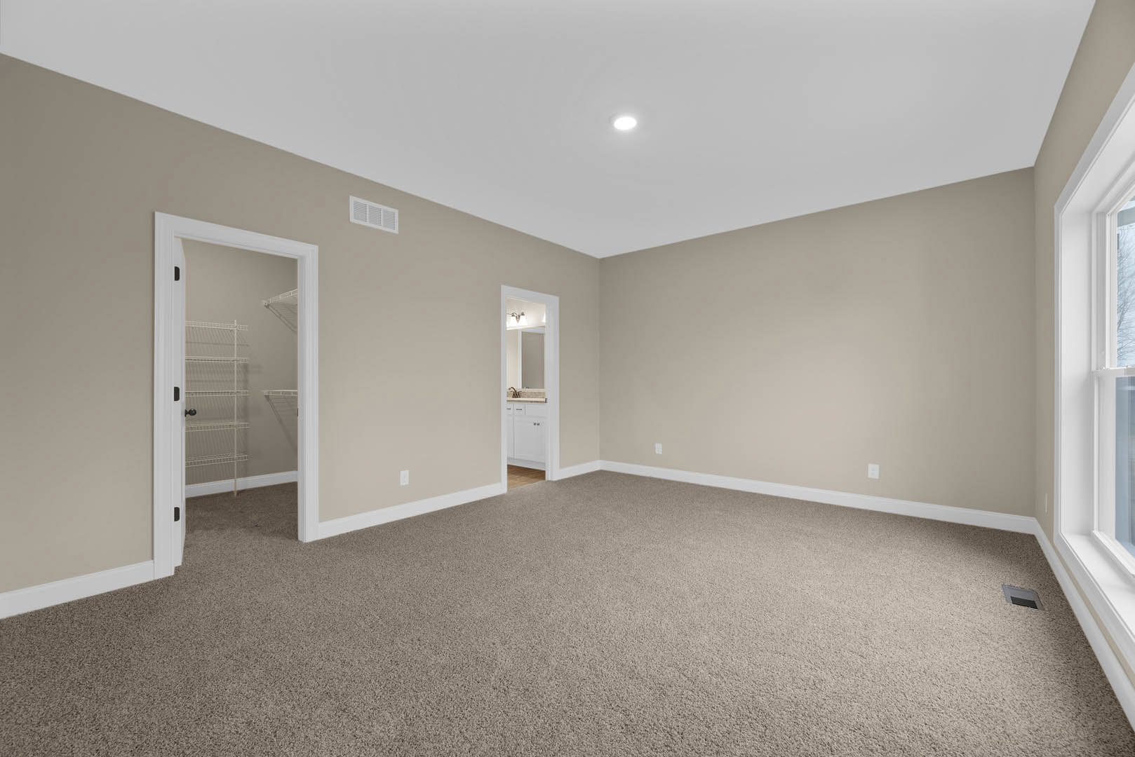 Beige-walled carpeted bedroom with open door, white closet shelving, black door knob, ceiling light, and partial view of bathroom sink and mirror