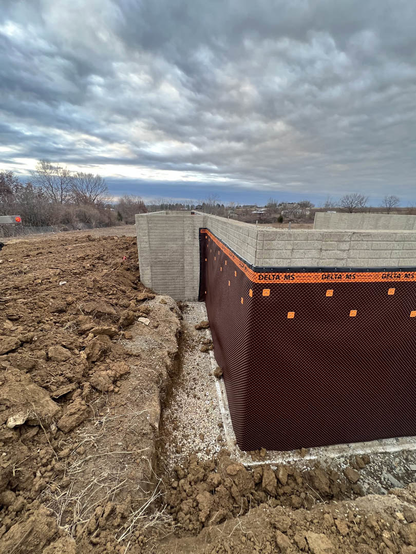 Grey brick wall under cloudy sky, bordered by dirt field and scattered construction materials, with trees in background.