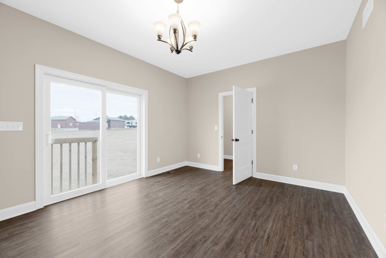 Bedroom with wood flooring and white trim, sliding glass door opening to balcony overlooking a field, white door with black knob, white wooden headboard, chandelier hanging from