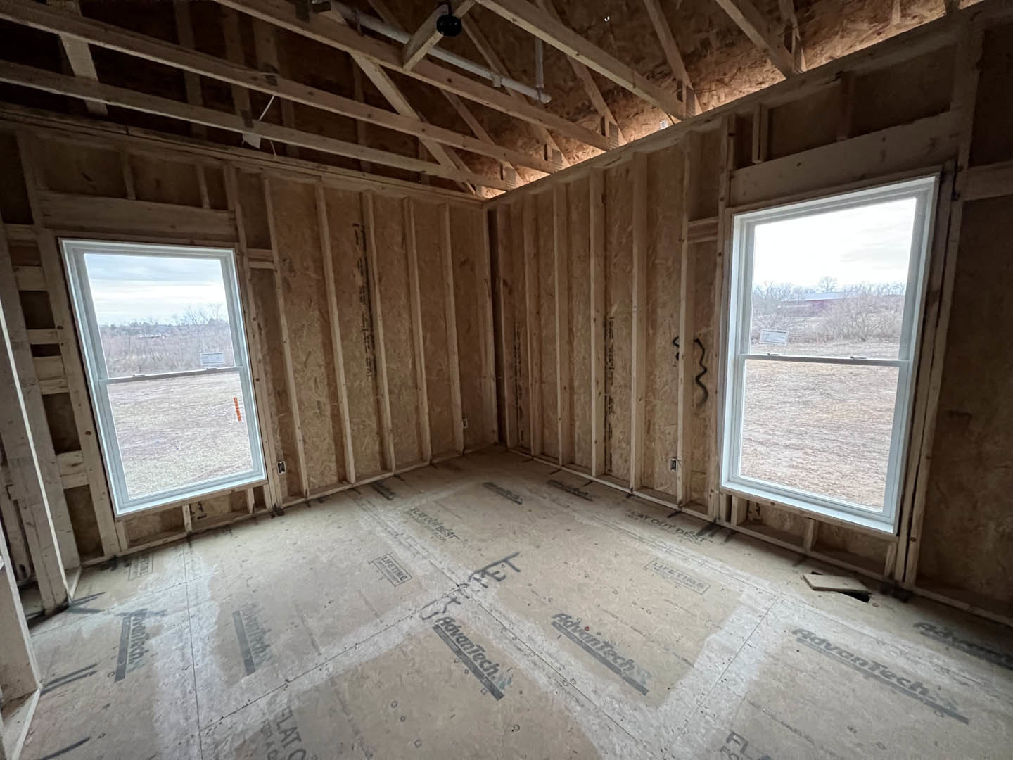 Spacious room featuring exposed wood ceiling beams, large windows overlooking a grassy field, neutral walls, and a dark floor accent