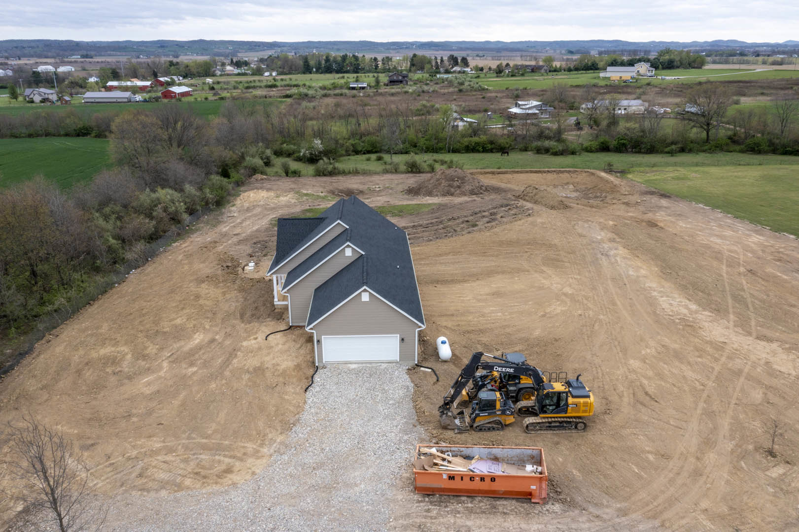 Partially built house with exposed framing, attached garage, construction tractor parked on dirt lot, leafless tree nearby, cardboard container and other construction materials