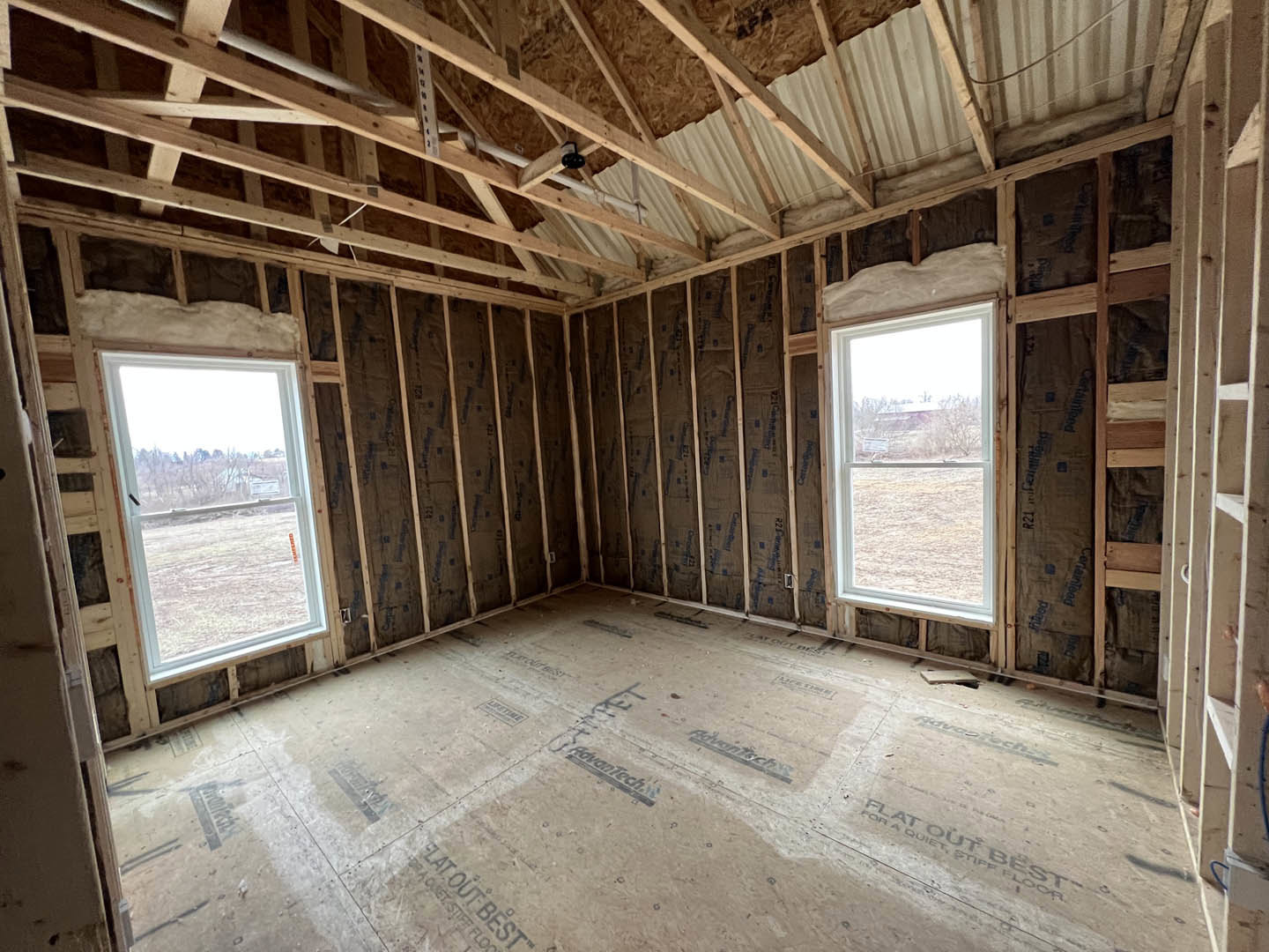 Living room with exposed wood ceiling beams, large windows overlooking a grassy field, wood-framed walls, and a white ceiling with brown trim.