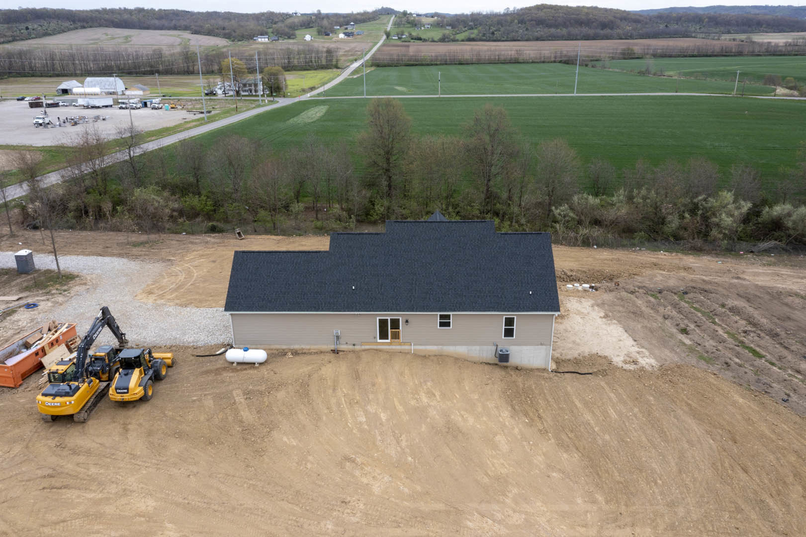 Partially built house with black roof, exposed framing, large window, construction tractor with crane and dumpster filled with wood, surrounded by field and trees under open sky