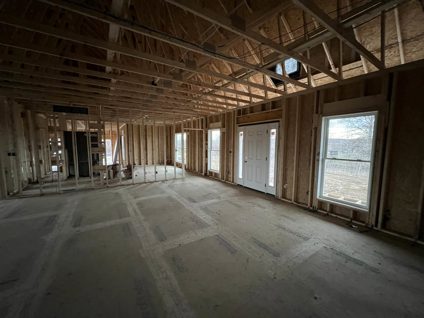 Wood-framed room with exposed ceiling beams, multiple windows, and a white door with glass panes, natural daylight illuminating unfinished floors.