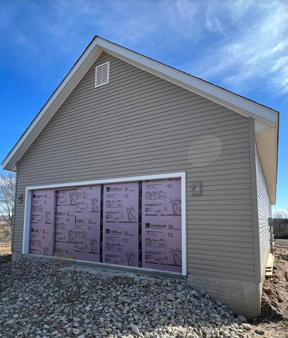 Grey-sided custom home with purple front and garage doors, white vent on exterior wall, pile of rocks near foundation, cloudy sky above, and shed visible in background.