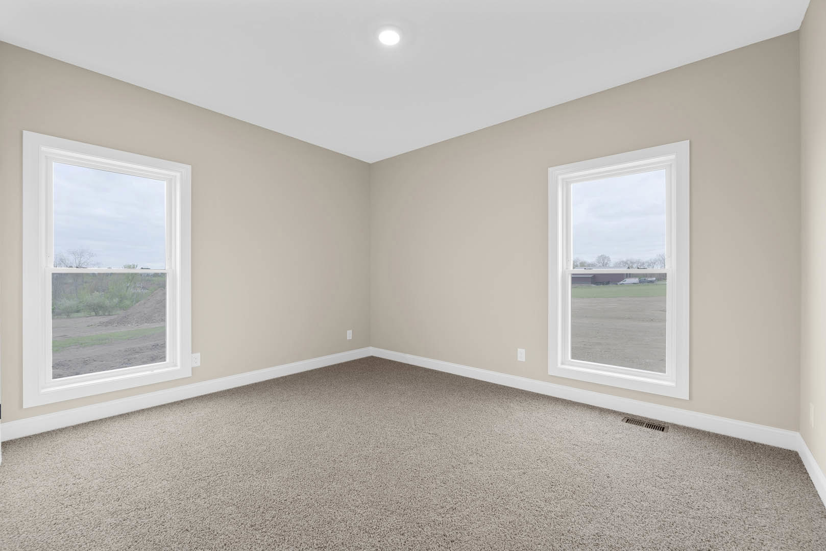 Carpeted room with two windows, white walls, ceiling light fixture, views of a grassy field with parked cars and a dirt field outside