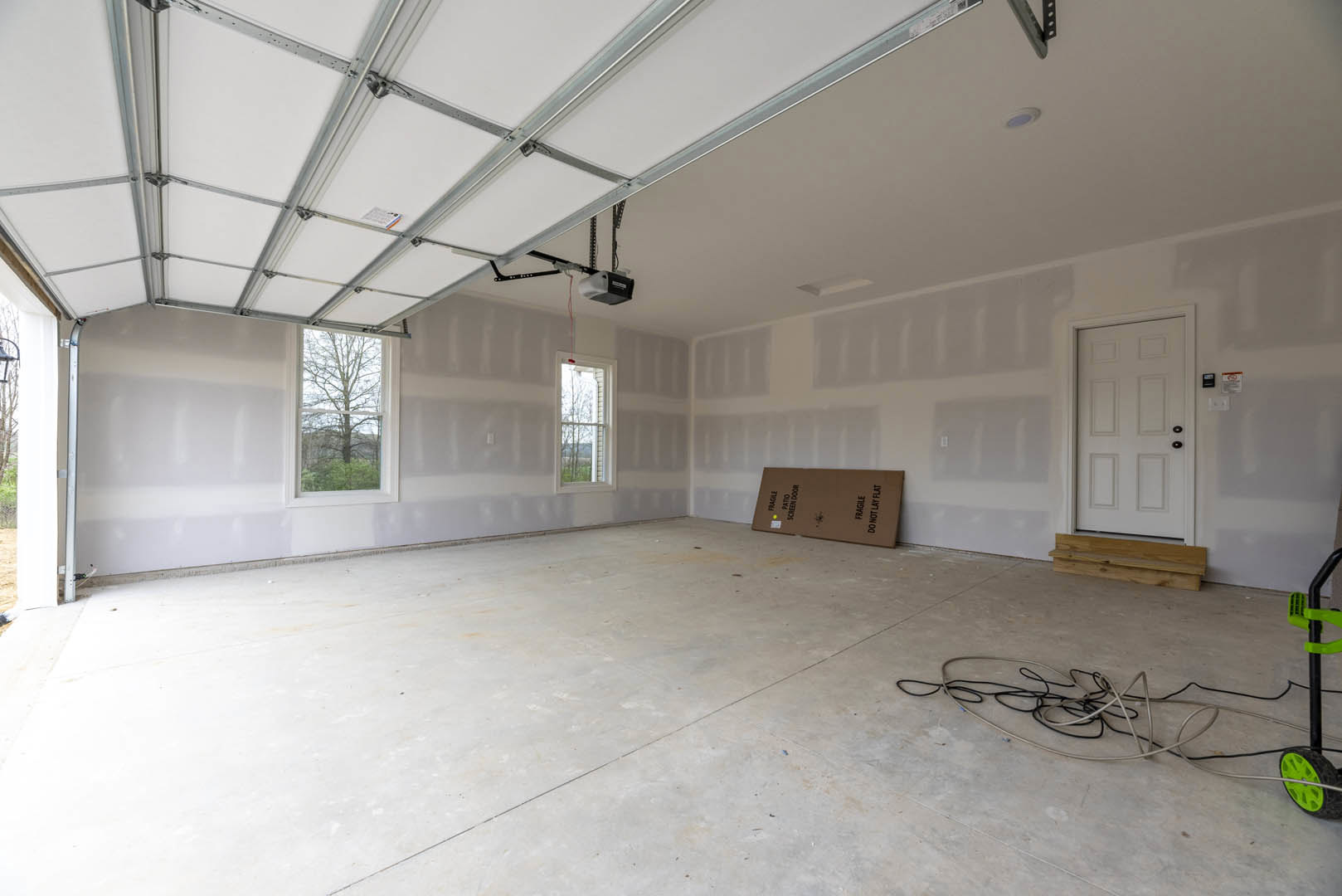 White plaster wall, black cable running along grey composite flooring, white door with black knobs, window showing leafy tree outside, ceiling-mounted projector, cardboard box near