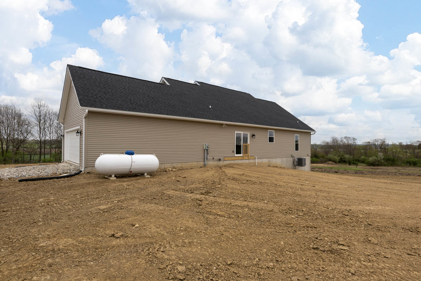 Single-story house with gray roof and beige siding, large cylindrical white tank with blue top positioned on dirt field in front, grassy area and trees in background under partly