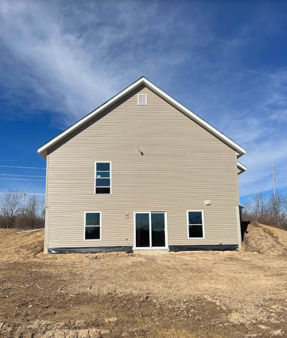 Partially built house with white framed windows and sliding glass door, light siding, dirt hill in foreground, blue sky, power lines, and historic Rockingham Meeting House visible