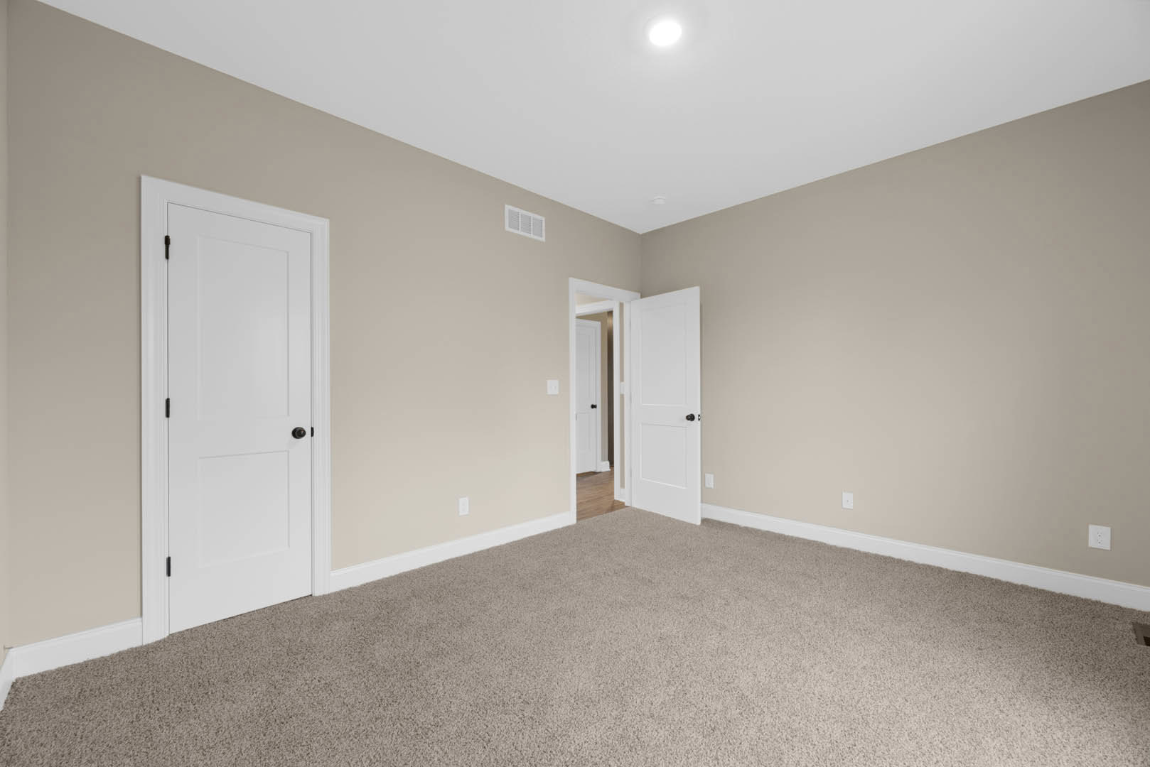 Beige-walled room with white paneled doors, black door knob, light gray carpet flooring, ceiling light fixture, and white wall vent