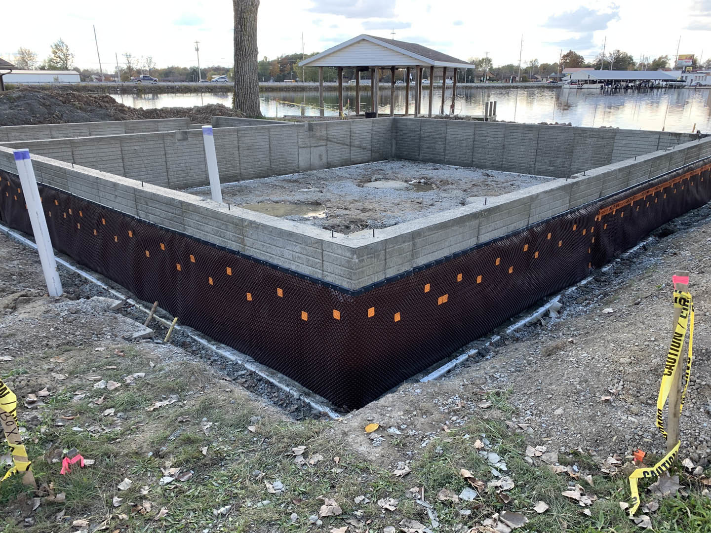 Concrete house foundation with wooden framing, surrounded by dirt and construction materials, yellow caution tape on a nearby pole, tree trunk and greenery in the background