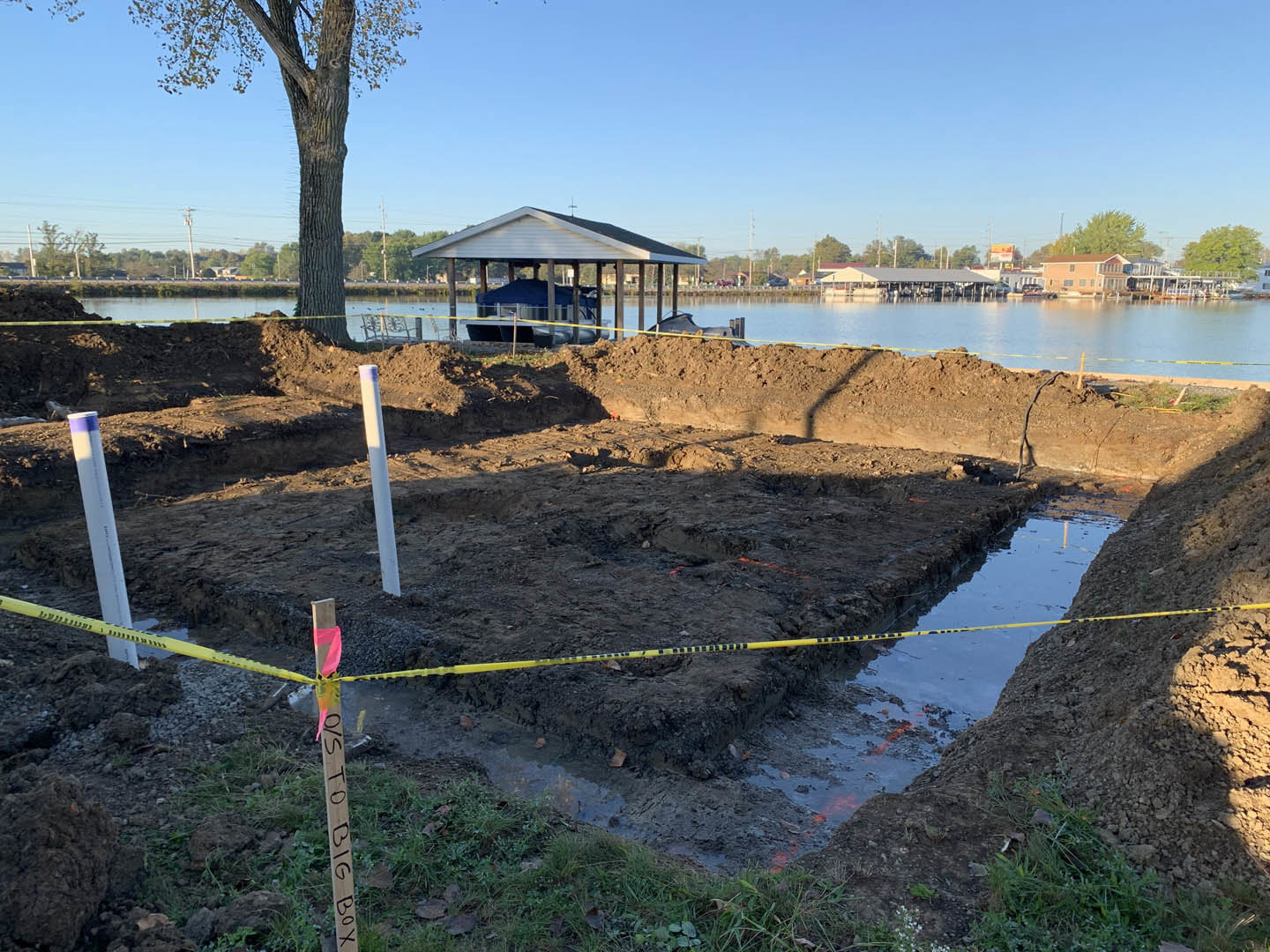 White house under construction with blue tarp, yellow caution tape on dirt, wooden post with markings, tree beside lake, clear blue sky