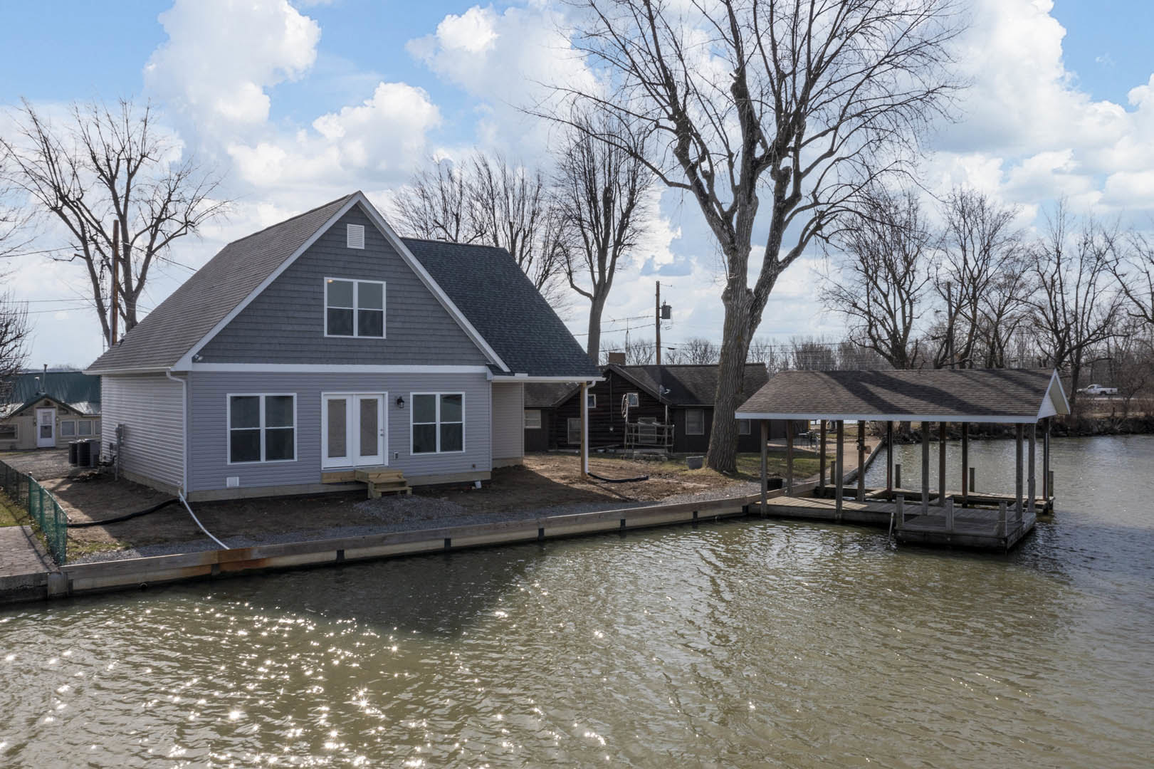 Two-story cottage with gray siding and multiple windows, situated beside a lake with a wooden dock extending over the water; mature trees and a small boat visible in the background