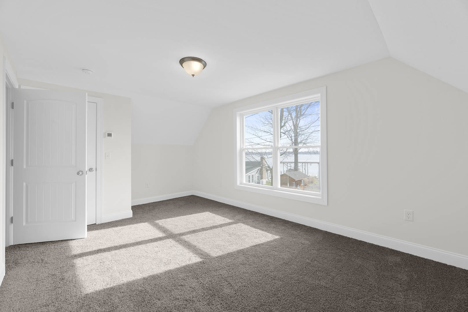 Carpeted room with white walls, large window showing trees and neighboring house, white door with silver knobs, ceiling light fixture with white shade