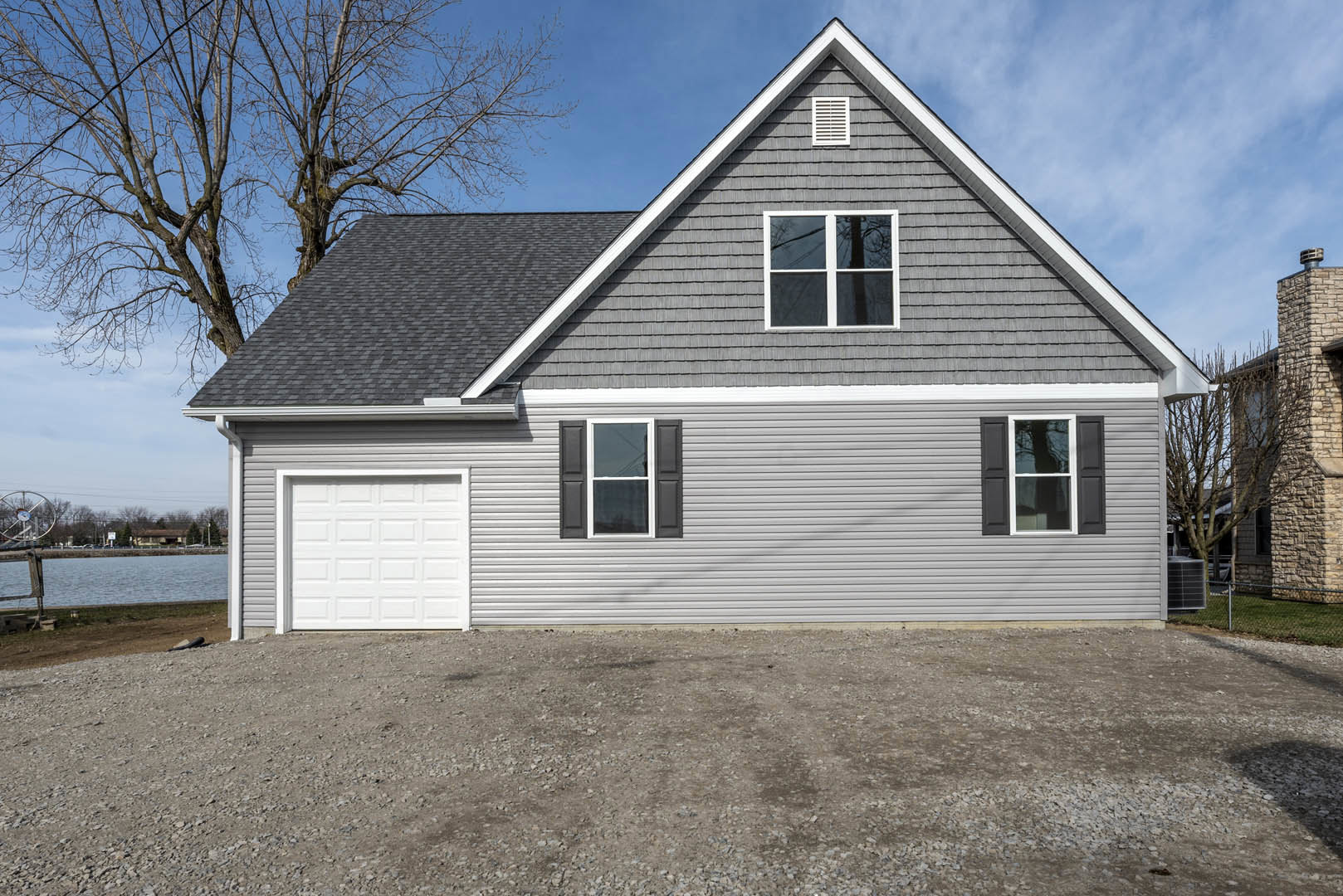 Two-story home with gray siding, white-framed windows, black shutters, attached garage with white door, mature tree in front yard, dirt driveway, cloudy sky overhead